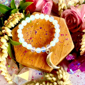 May include: A white opal beaded bracelet with a silver crescent moon charm. The bracelet is on a wooden surface with pink and white flowers in the background.