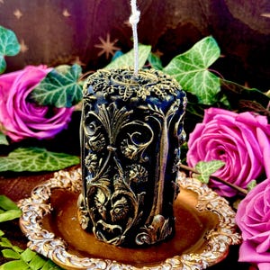 May include: A black and gold candle with an ornate design. The candle is sitting on a gold tray.
