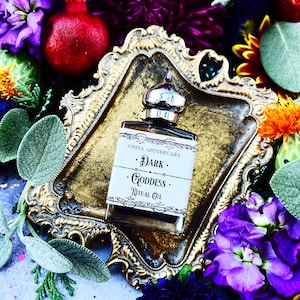 May include: A small glass bottle with a gold lid and a label that reads "Anita Apothecary - Dark Goddess - Ritual Oil" sits on a gold tray surrounded by purple, blue, and yellow flowers.