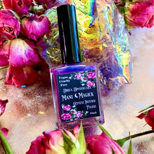 May include: A square bottle of purple nail polish with a black cap. The label reads "Anita Apothecary Mani Magick Crystal Infused Polish" with floral accents. The words "Vegan Cruelty Free" are also on the label. The bottle is surrounded by dried roses.