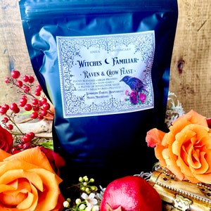 May include: A black bag labeled "Witches Familiar Raven & Crow Feast" with a decorative label featuring a raven and floral accents. The bag is surrounded by orange roses, red berries, and a pomegranate.