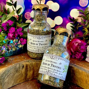 May include: Two glass bottles filled with dried herbs, labeled "Kitchen Witchery Herbs de Provence." The bottles have gold lids and are sitting on a wooden surface with colorful glitter scattered around them.