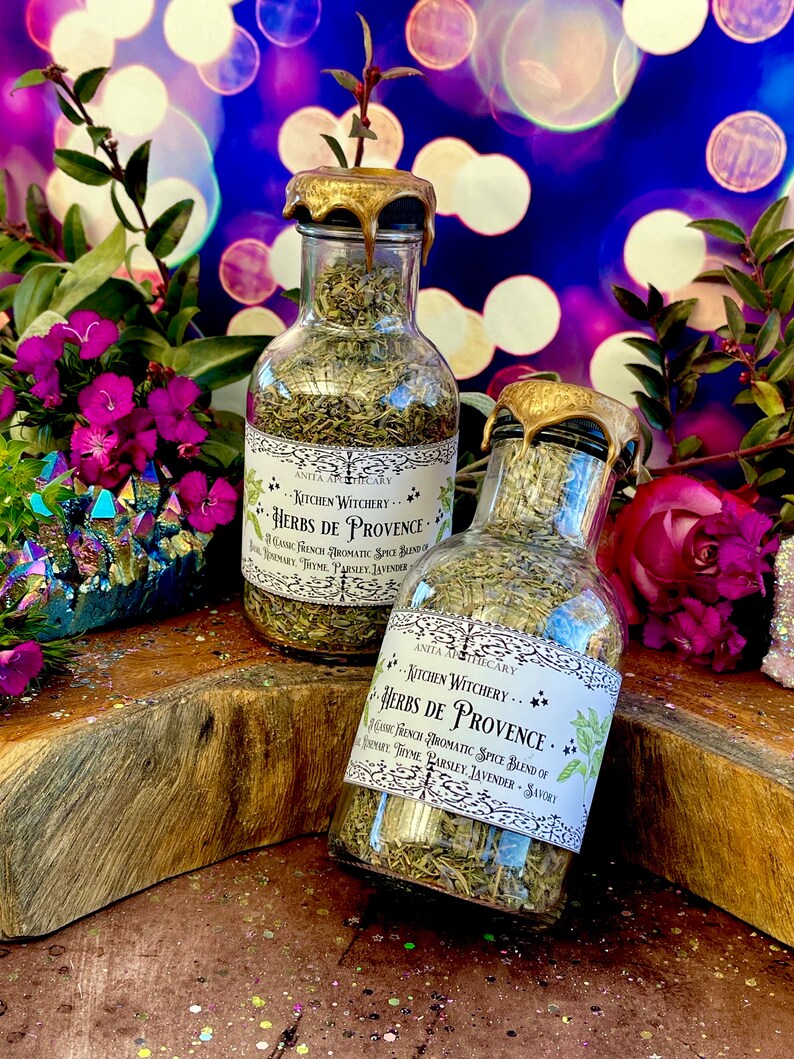 May include: Two glass bottles filled with dried herbs, labeled "Kitchen Witchery Herbs de Provence." The bottles have gold lids and are sitting on a wooden surface with colorful glitter scattered around them.