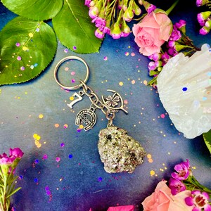 Pode incluir: Um chaveiro de prata com uma ferradura, um trevo de quatro folhas e um pequeno cristal de rocha. Os pingentes est&atilde;o presos a um anel de chave de prata. O chaveiro &eacute; rodeado por flores cor-de-rosa e glitter iridescente.