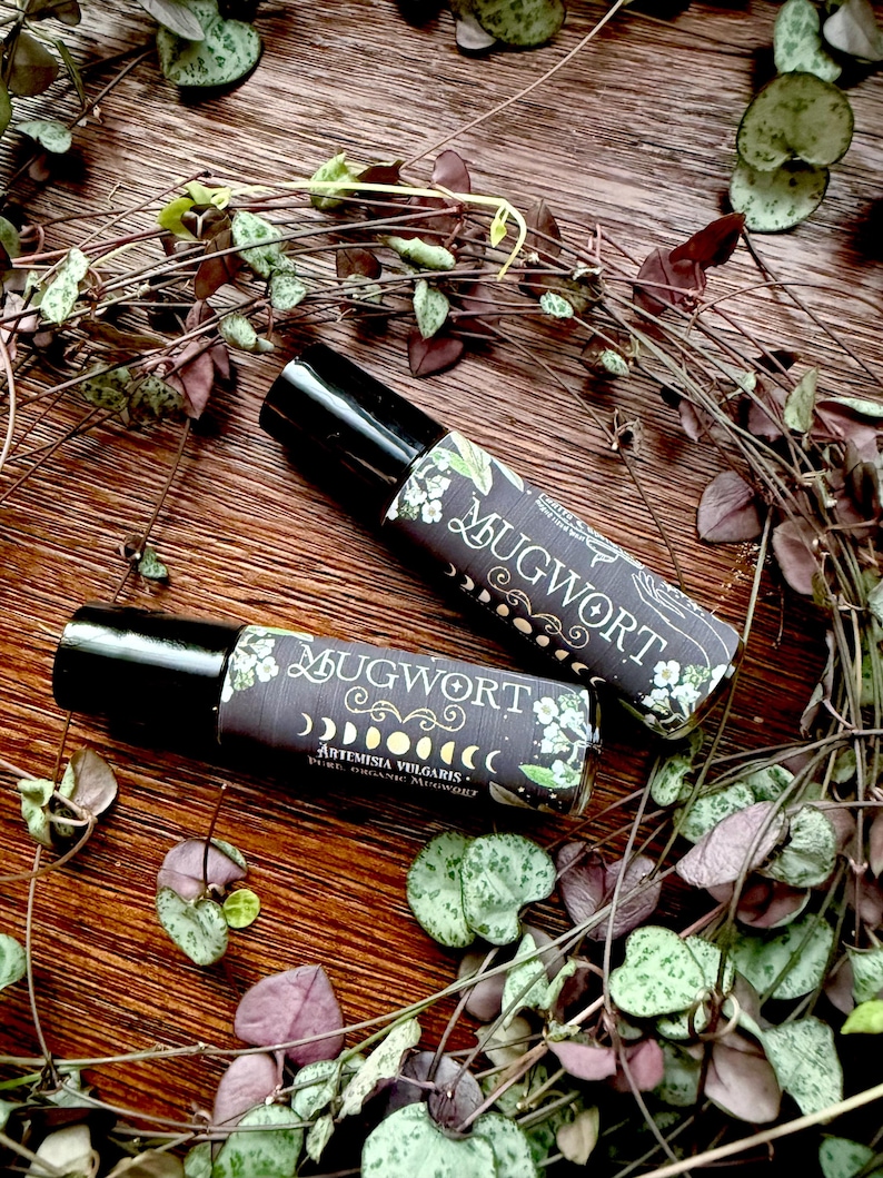 May include: Two small glass bottles with black roll-on tops. Both bottles have a black and white label with the text "Mugwort" and "Artemisia Vulgaris - Pure Organic Mugwort". The bottles are surrounded by green and purple leaves.
