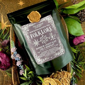 May include: A dark green package of "Folklore" hand-blended botanical teas, sealed with a gold wax stamp. The label features ornate text and botanical illustrations. The package is surrounded by flowers and greenery, creating a natural, earthy aesthetic.