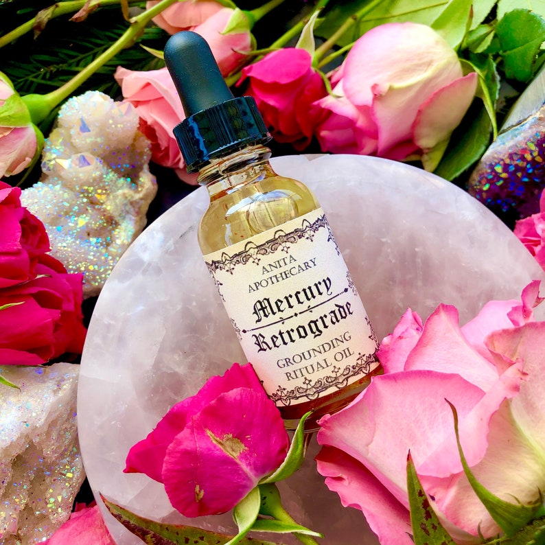 Puede incluir: Una peque&ntilde;a botella de vidrio con un gotero llena de un l&iacute;quido dorado. La botella tiene una etiqueta que dice "Anita Apothecary Mercury Retrograde Grounding Ritual Oil". La botella est&aacute; rodeada de rosas rosadas y cristales blancos.