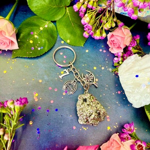 Pode incluir: Um chaveiro de prata com uma ferradura, um trevo de quatro folhas e uma pequena pedra dourada brilhante. O chaveiro &eacute; rodeado por flores rosa e roxas e glitter.