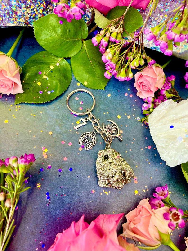 Pode incluir: Um chaveiro de prata com uma ferradura, um trevo de quatro folhas e uma pequena pedra dourada brilhante. O chaveiro &eacute; rodeado por flores rosa e roxas e glitter.