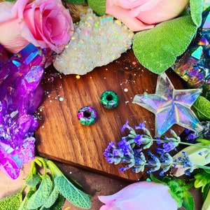 May include: Two green and pink speckled round beads on a wooden surface with flowers and crystals in the background.