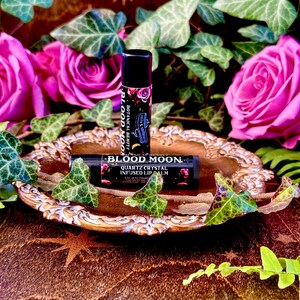 May include: Two black lip balm tubes with the words "Blood Moon" and "Quartz Crystal Infused Lip Balm" on them. The tubes are on a gold-colored tray with green leaves and pink roses around them.