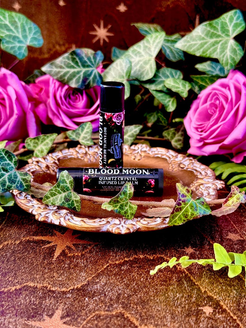 May include: Two black lip balm tubes with the words "Blood Moon" and "Quartz Crystal Infused Lip Balm" on them. The tubes are on a gold-colored tray with green leaves and pink roses around them.