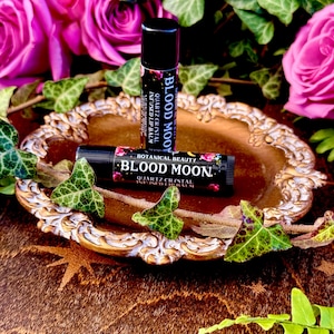 May include: Two black lip balm tubes with gold lettering on a gold decorative plate. The lip balm tubes are labeled "Blood Moon" and "Botanical Beauty Blood Moon." The lip balm tubes are surrounded by green leaves and pink roses.