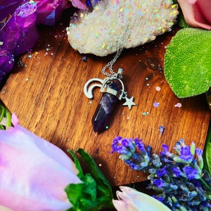 May include: A silver necklace with a purple amethyst crystal pendant, a crescent moon charm, and a star charm. The necklace is displayed on a wooden surface with pink roses, lavender, and a white crystal cluster.