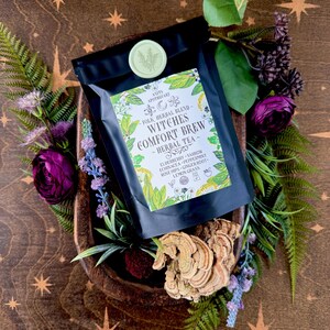 May include: A black bag of "Witches Comfort Brew" herbal tea, surrounded by greenery and purple flowers. The tea bag has a green and white label with the Anita Apothecary logo and a list of ingredients. The background is a wooden bowl with a starburst pattern.