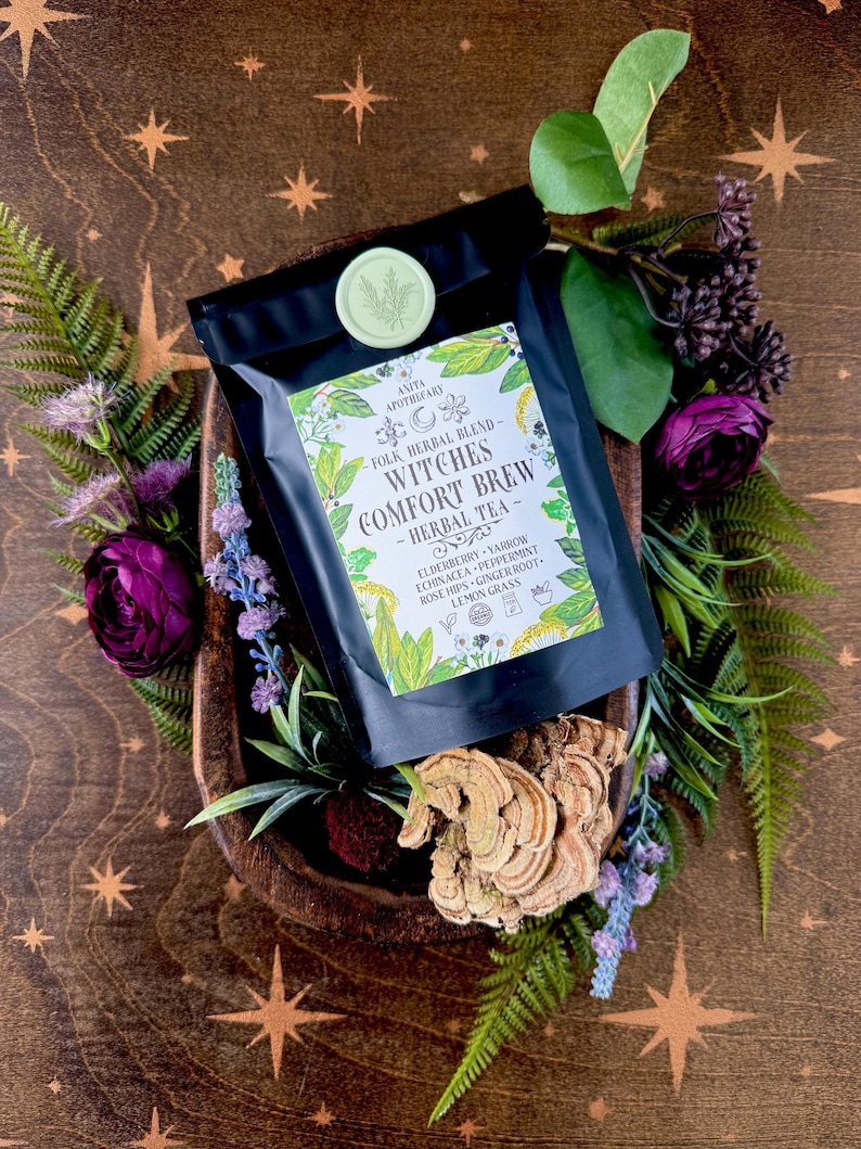 May include: A black bag of "Witches Comfort Brew" herbal tea, surrounded by greenery and purple flowers. The tea bag has a green and white label with the Anita Apothecary logo and a list of ingredients. The background is a wooden bowl with a starburst pattern.