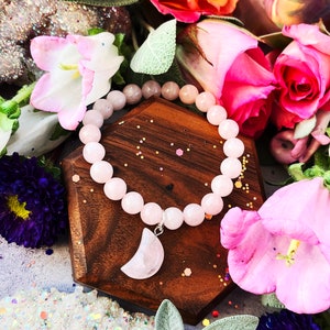 May include: A pink rose quartz bracelet with a crescent moon charm. The bracelet is made of round beads and is displayed on a wooden stand with pink and white flowers.