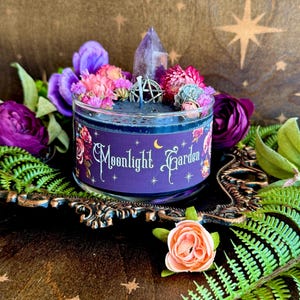May include: A round candle in a clear glass jar with dark wax, topped with a purple crystal point and a silver pentacle charm. The label features the text Moonlight Garden with floral illustrations, surrounded by dried flowers and green ferns.