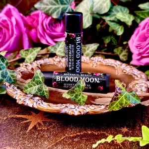 May include: Two tubes of lip balm with the words "Blood Moon" and "Quartz Crystal Infused Lip Balm" on the labels. The tubes are on a gold-colored tray with a star design.