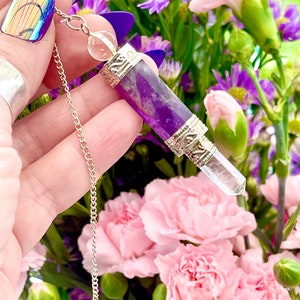 May include: Amethyst pendulum with a clear crystal point and silver accents. The pendulum is hanging from a silver chain and is surrounded by purple and pink flowers.