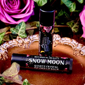 May include: Two black lip balm tubes with gold lettering. The top tube says "Botanical Beauty" and "Snow Moon" with a crescent moon and stars. The bottom tube says "Quartz Crystal Infused Lip Balm".