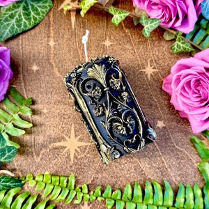 May include: A black and gold decorative pillar candle with an intricate floral design. The candle is surrounded by green foliage and pink roses.