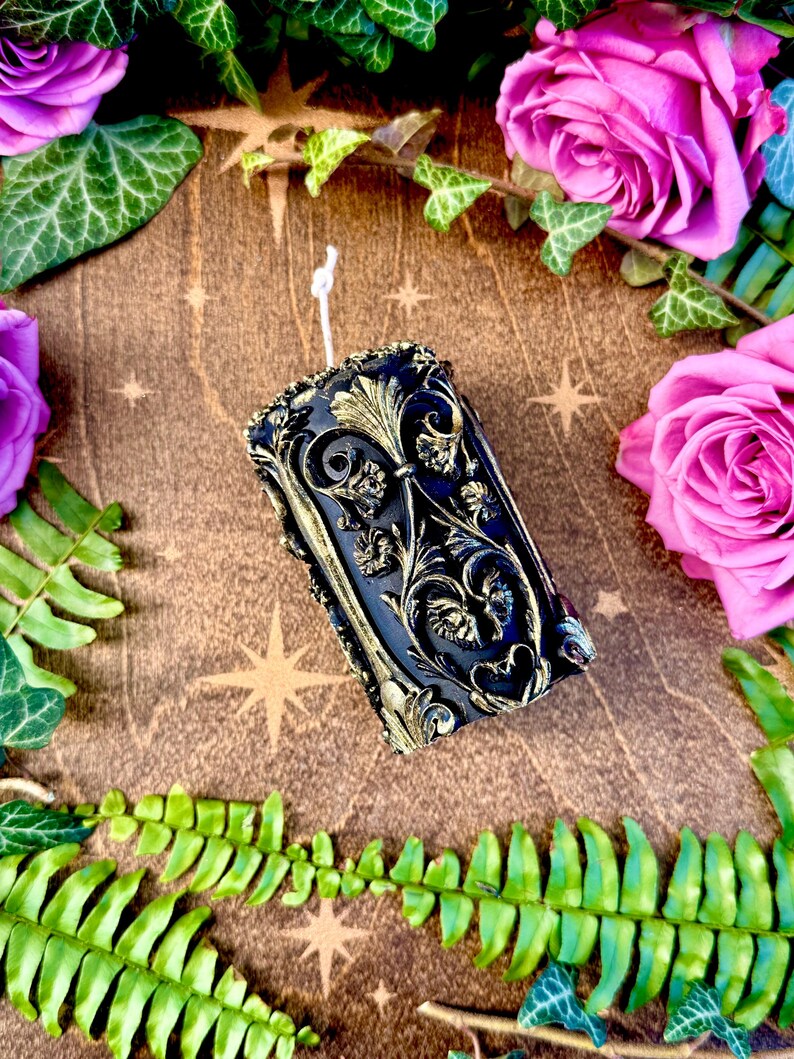 May include: A black and gold decorative pillar candle with an intricate floral design. The candle is surrounded by green foliage and pink roses.