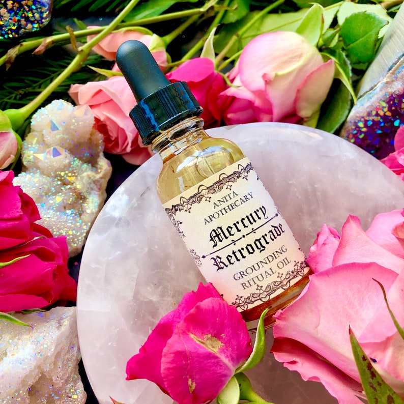 Puede incluir: Una peque&ntilde;a botella de vidrio con un gotero, llena de un l&iacute;quido transparente. La botella tiene una etiqueta que dice "Anita Apothecary Mercury Retrograde Grounding Ritual Oil". La botella est&aacute; rodeada de rosas rosadas y cristales blancos.