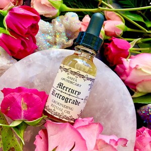 Puede incluir: Una peque&ntilde;a botella de vidrio con un gotero llena de un l&iacute;quido transparente. La botella tiene una etiqueta blanca con texto negro que dice "Anita Apothecary Mercury Retrograde Grounding Ritual Oil". La botella est&aacute; rodeada de rosas rosadas y un cristal blanco.