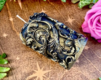May include: A black and gold decorative candle with an ornate design. The candle is on a wooden surface with green foliage.