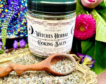 May include: A glass jar of "Witches Herbal Cooking Salts" with a black lid and a label. The label reads "Anita Apothecary". A wooden spoon rests on a decorative tray filled with the salt blend. The image is surrounded by flowers.