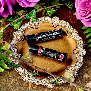 May include: Two small black tubes of lip balm with floral designs and the text "Botanical Beauty Blood Moon Quartz Crystal Infused Lip Balm" on a gold decorative tray with pink roses and green leaves.