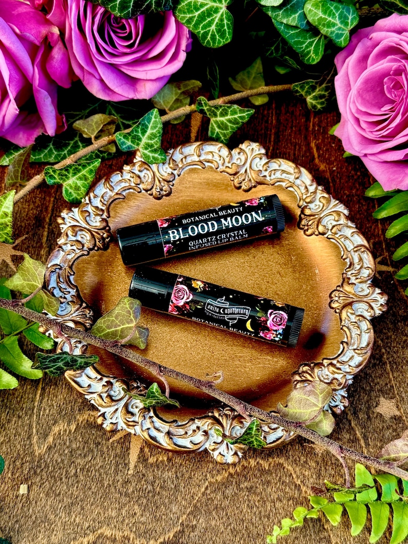 May include: Two small black tubes of lip balm with floral designs and the text "Botanical Beauty Blood Moon Quartz Crystal Infused Lip Balm" on a gold decorative tray with pink roses and green leaves.