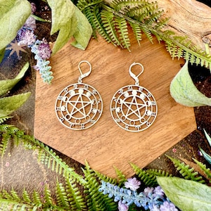 May include: A pair of silver-colored earrings with a circular design featuring a pentagram and astrological symbols. The earrings are set against a wooden background, surrounded by green foliage and purple flowers.