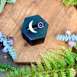May include: A silver ring featuring a crescent moon and a purple gemstone. The ring is displayed on a black hexagonal stand, surrounded by greenery and flowers. The ring's design is simple and elegant.