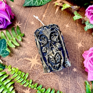 May include: A black pillar candle with a gold ornate design. The candle is surrounded by green leaves and pink flowers.