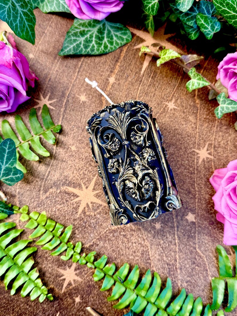 May include: A black pillar candle with a gold ornate design. The candle is surrounded by green leaves and pink flowers.