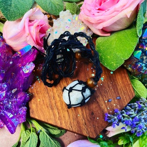 May include: A white crystal stone wrapped in black cord with wooden beads, displayed on a wooden hexagon-shaped surface surrounded by flowers and crystals.