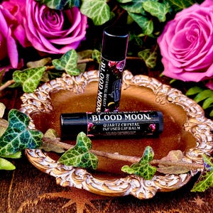 May include: Two black tubes of Blood Moon Quartz Crystal Infused Lip Balm on a gold decorative tray with pink roses and green ivy.