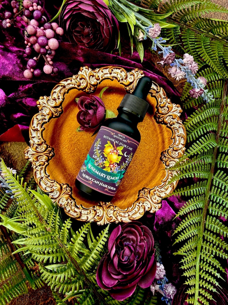 Pode incluir: Um frasco preto de s&eacute;rum capilar marroquino Rosemary Lemon da Anita Apothecary com conta-gotas, colocado numa bandeja dourada ornamentada. A bandeja &eacute; rodeada por veludo roxo, flores e fetos verdes. O r&oacute;tulo apresenta ilustra&ccedil;&otilde;es bot&acirc;nicas.