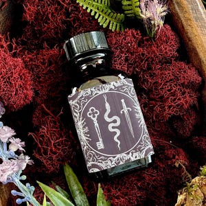 May include: A dark glass bottle with a black cap and a label featuring a key, snake, and sword design. The bottle sits on a bed of red moss, with green and purple foliage accents. The overall aesthetic is mystical and earthy.
