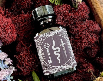 Può includere: Una bottiglia di vetro scuro con tappo nero ed etichetta con chiave, serpente e spada. La bottiglia poggia su muschio rosso, con accenti di fogliame verde e viola. L'estetica generale è mistica e terrena.