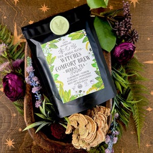 May include: A black bag of "Witches Comfort Brew" herbal tea, with a green and white label, sits in a wooden bowl surrounded by flowers and foliage. The label lists ingredients like elderberry and peppermint. A green wax seal is visible.