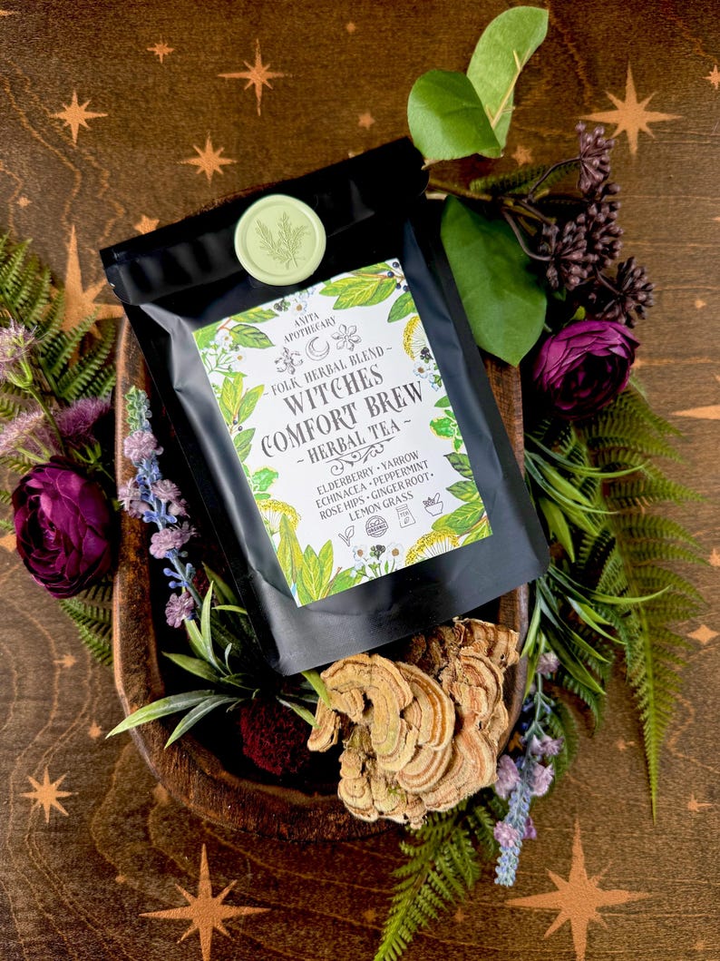 May include: A black bag of "Witches Comfort Brew" herbal tea, with a green and white label, sits in a wooden bowl surrounded by flowers and foliage. The label lists ingredients like elderberry and peppermint. A green wax seal is visible.