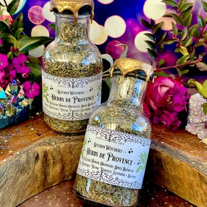 May include: Two glass bottles with gold lids filled with dried herbs. The labels read "Kitchen Witchery... Herbs de Provence" and list the ingredients as a classic French aromatic spice blend of rosemary, thyme, parsley, lavender, and savory.