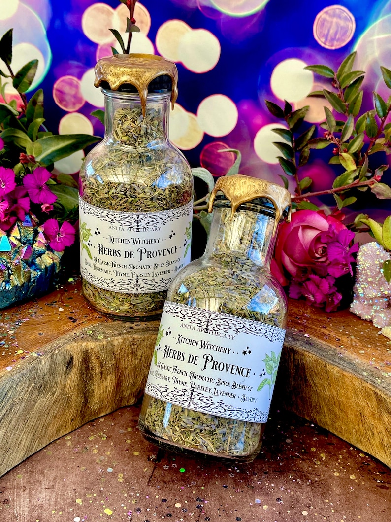 May include: Two glass bottles with gold lids filled with dried herbs. The labels read "Kitchen Witchery... Herbs de Provence" and list the ingredients as a classic French aromatic spice blend of rosemary, thyme, parsley, lavender, and savory.