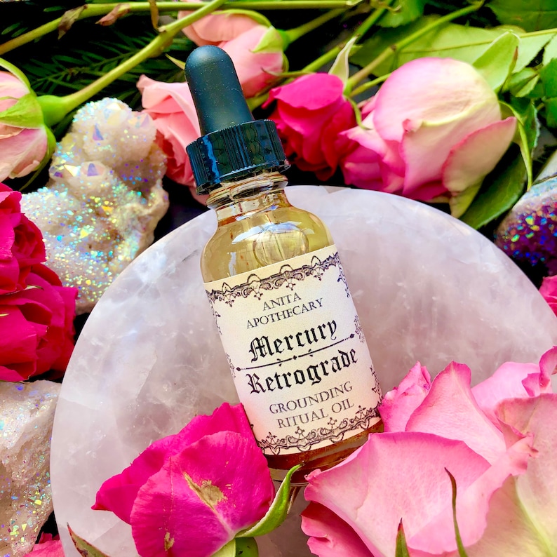 Puede incluir: Una peque&ntilde;a botella de vidrio con un gotero llena de un l&iacute;quido dorado. La etiqueta dice "Anita Apothecary Mercury Retrograde Grounding Ritual Oil". La botella est&aacute; rodeada de rosas rosadas y un cristal blanco.