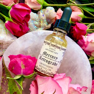 Puede incluir: Una peque&ntilde;a botella de vidrio con un gotero llena de un l&iacute;quido dorado. La etiqueta dice "Anita Apothecary Mercury Retrograde Grounding Ritual Oil". La botella est&aacute; rodeada de rosas rosas y un cristal blanco.