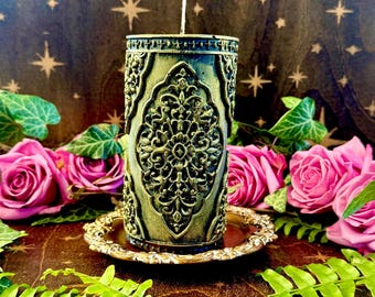 May include: A decorative candle with an ornate, embossed design in shades of green and gold, resting on a gold-colored saucer. The candle is surrounded by pink roses and green foliage, creating a mystical and elegant aesthetic.