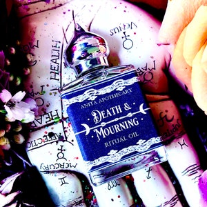 Pu&ograve; includere: Una bottiglia di vetro blu con un tappo argentato e un'etichetta bianca che recita "Anita Apothecary Death & Mourning Ritual Oil". La bottiglia &egrave; appoggiata su una superficie con uno sfondo colorato.